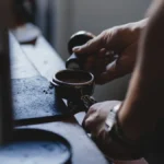 Barista uses tool to scrape crema from espresso shot in clean coffee shop setting.