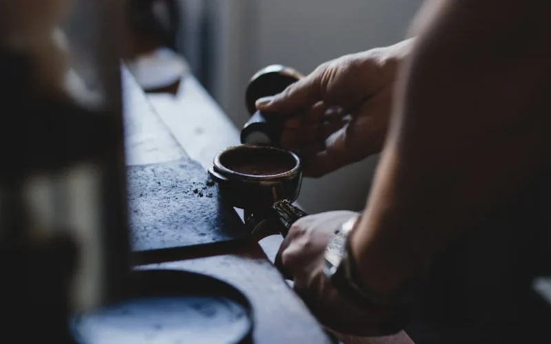 Barista uses tool to scrape crema from espresso shot in clean coffee shop setting.