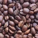 Coffee beans spilling from a conical mill into a dark wooden tray with steam rising nearby.