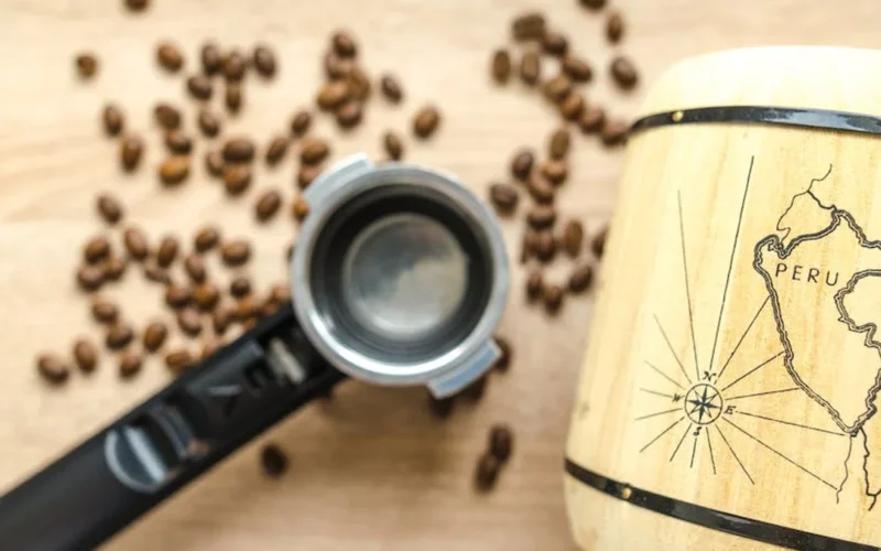 Green coffee beans spilling from a glass jar on a wooden table with steam rising from a nearby roaster.