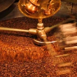 Dark roast coffee beans spilling from a drum roaster with steam rising from a kettle on a wooden counter.