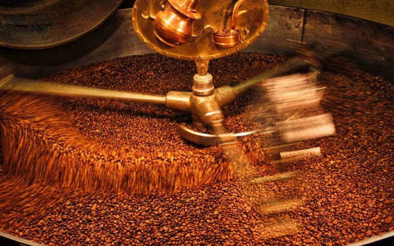 Dark roast coffee beans spilling from a drum roaster with steam rising from a kettle on a wooden counter.