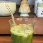 A frosted glass matcha latte with steam, wooden table, and ceramic tea set under golden light.
