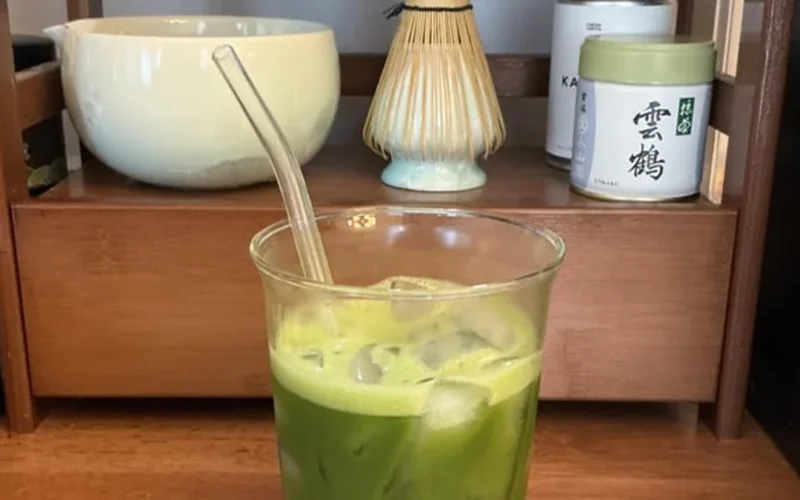 A frosted glass matcha latte with steam, wooden table, and ceramic tea set under golden light.