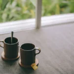 A steaming coffee cup on a wooden table with a plant and natural light.