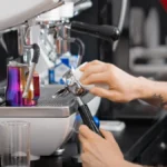 Barista tampes espresso with handheld auto tamper tool under warm lighting.