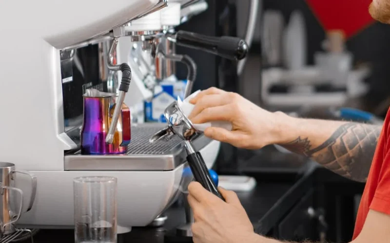 Barista tampes espresso with handheld auto tamper tool under warm lighting.