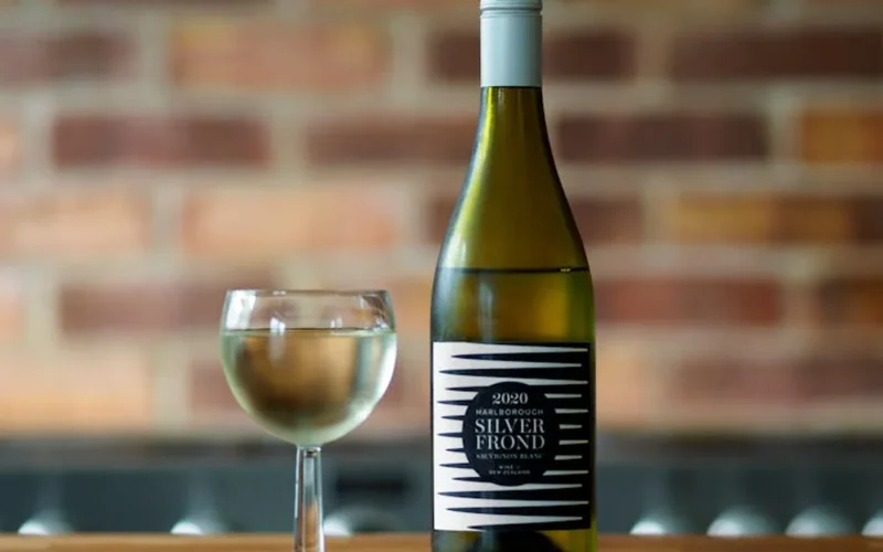 Orange wine bottle with disclaimer label next to a glass of sparkling wine on a wooden table under natural light.