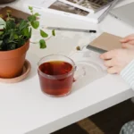 Tea taster examining tea liquid under magnifying glass with tea wheel chart and notebook on wooden table.
