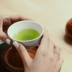 Bright green matcha powder in a bowl beside traditional dried sencha tea leaves.