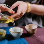Hands pouring tea from a traditional Chinese clay teapot into a glass vessel.