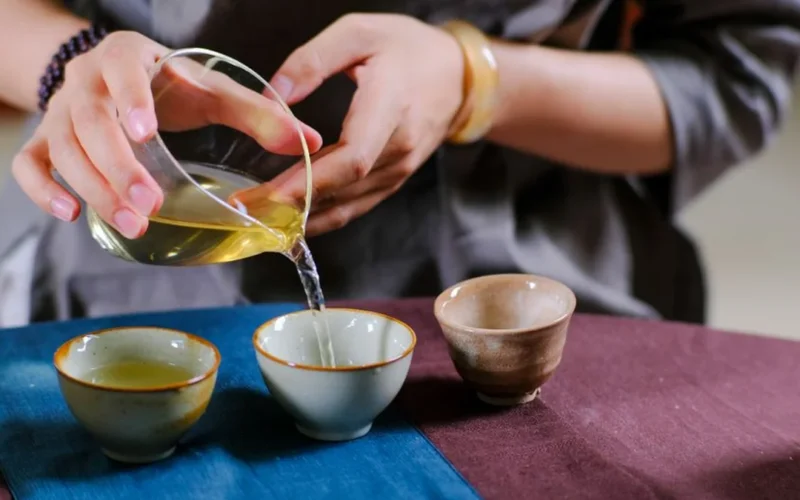 Hands pouring tea from a traditional Chinese clay teapot into a glass vessel.