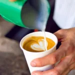 Close up of a barista pouring milk to create latte art in a ceramic cup.