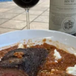 A bottle of vintage red wine and a glass served with grilled steak on wood.
