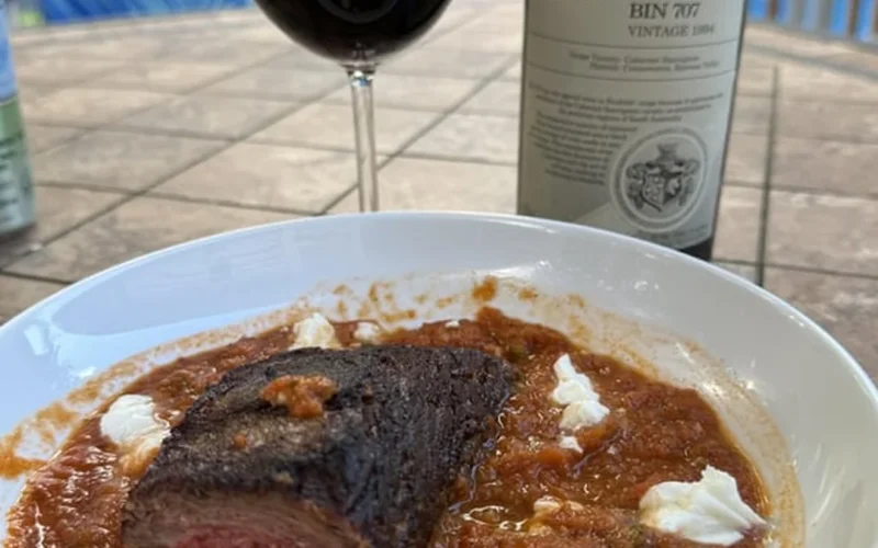A bottle of vintage red wine and a glass served with grilled steak on wood.