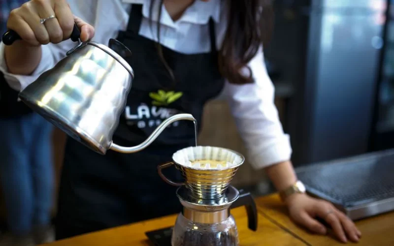 A steaming glass carafe of black drip coffee next to a ceramic mug on wood.