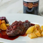 A glass of red Cabernet Sauvignon next to a steak dinner on a wooden table.