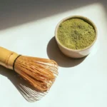 Bright green matcha powder and bamboo whisk on a dark stone surface.