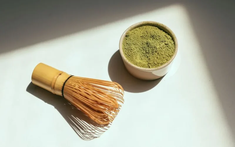 Bright green matcha powder and bamboo whisk on a dark stone surface.