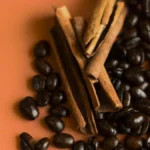 Close-up of roasted coffee beans, cacao pieces, and cinnamon sticks on a wooden surface.