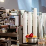 A busy cafe counter with disposable coffee cups and a ceramic mug in soft morning light.