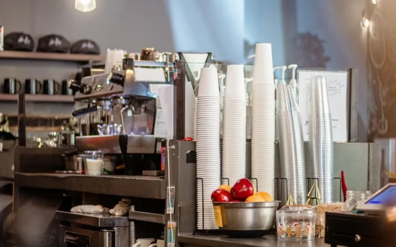 A busy cafe counter with disposable coffee cups and a ceramic mug in soft morning light.