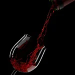 A glass of deep red Bordeaux wine next to a bottle on a dark textured surface.