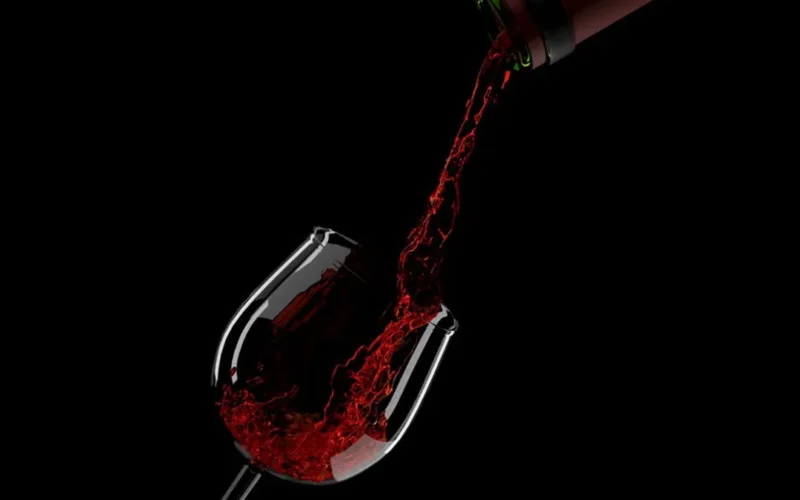 A glass of deep red Bordeaux wine next to a bottle on a dark textured surface.