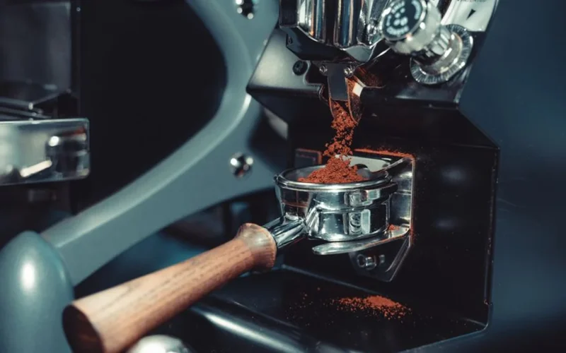 Close-up of a professional Macap coffee grinder dispensing grounds into a portafilter in a busy cafe.