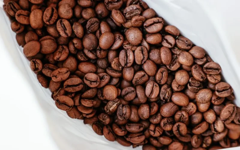 Dark roasted coffee beans scattered next to a black coffee bag on a dark surface.