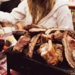 A glass of red wine next to pork cutlets and roasted carrots on a wooden table.