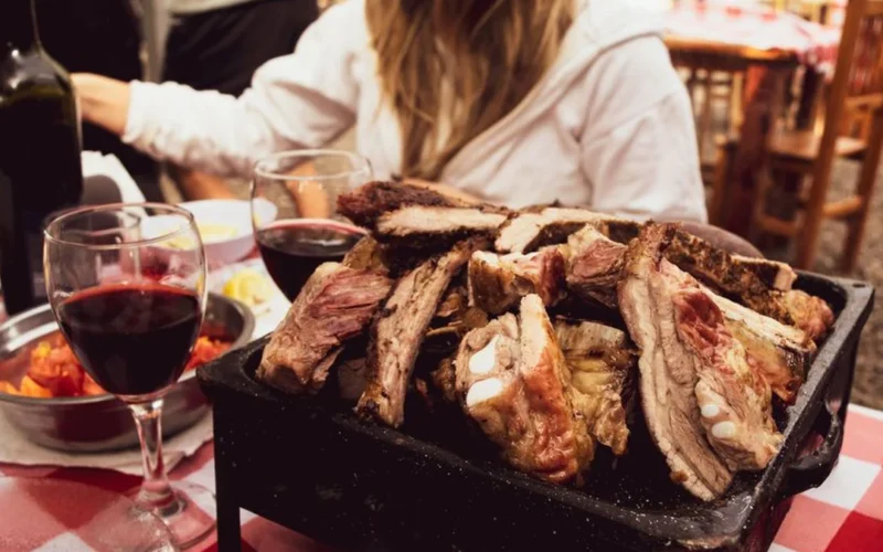 A glass of red wine next to pork cutlets and roasted carrots on a wooden table.
