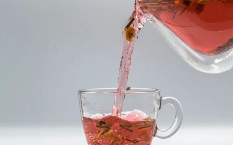 A glass teapot with amber tea next to a clear glass of water on wood.