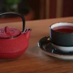 A cup of dark black tea resting on a wooden surface with dried tea leaves.