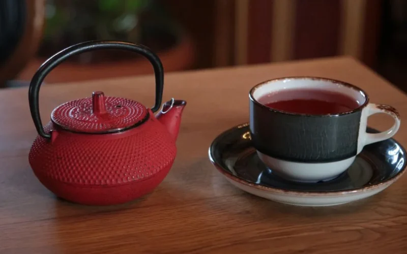 A cup of dark black tea resting on a wooden surface with dried tea leaves.