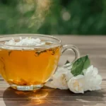 A glass cup of jasmine green tea surrounded by dried jasmine flower blossoms on stone.