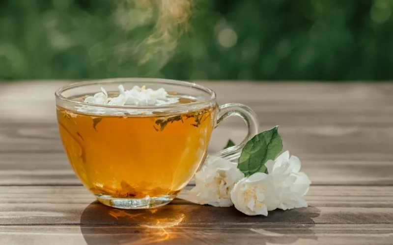 A glass cup of jasmine green tea surrounded by dried jasmine flower blossoms on stone.