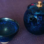 A dark Tenmoku ceramic bowl filled with light green tea on a wooden surface.