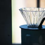 A glass V60 pour over dripper sitting on a wooden surface with coffee grounds inside.
