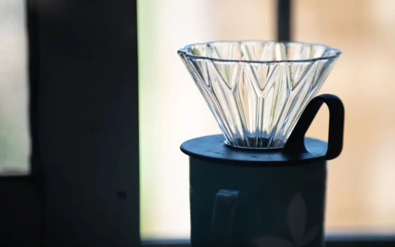 A glass V60 pour over dripper sitting on a wooden surface with coffee grounds inside.