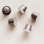 Shiny aluminum coffee capsules and roasted coffee beans on a dark stone surface.
