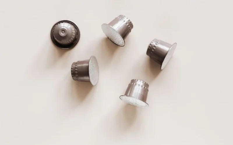 Shiny aluminum coffee capsules and roasted coffee beans on a dark stone surface.