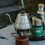 Overhead view of an insulated mug, immersion heater, and coffee beans on a flat surface.