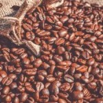 Roasted coffee beans spilling from a burlap sack next to a ceramic mug on wood.