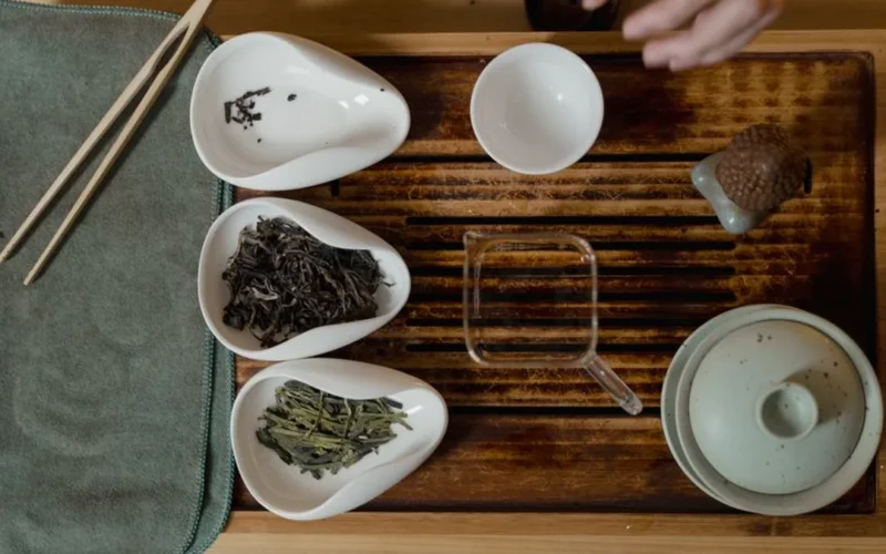 Dark loose puerh tea leaves scattered next to a white ceramic gaiwan on stone.