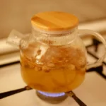 A glass kettle of golden Karak chai with cinnamon sticks and cardamom pods on a wooden table.
